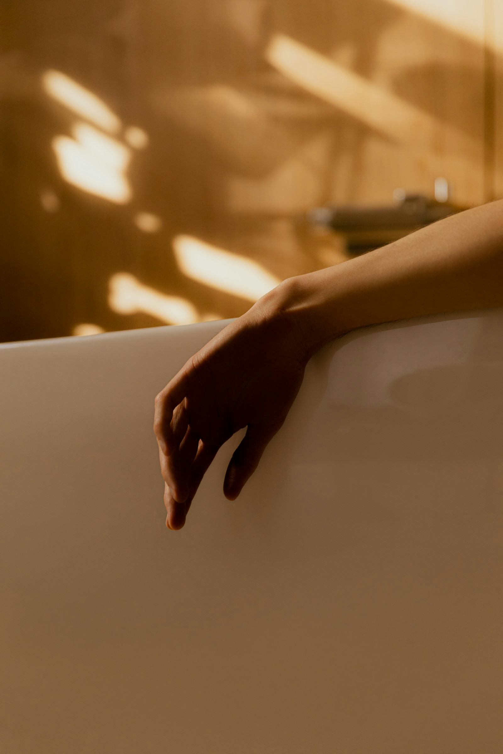 Close-up of a hand resting on the edge of a modern white bathtub, with warm lighting reflecting off the surface.