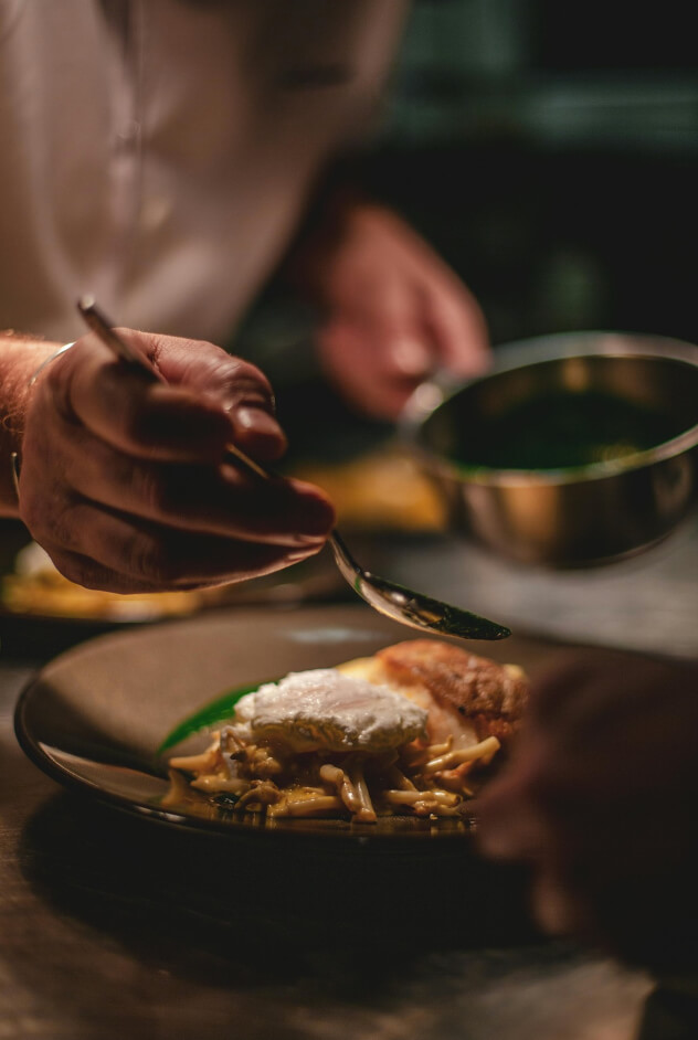 Close-up of a chef garnishing a dish with a spoon, preparing a meal in a restaurant kitchen, focusing on the details of the food.