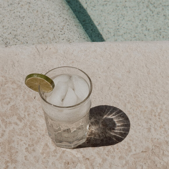 Refreshing glass of water with ice cubes and a lime slice, placed on a stone surface by the pool, with a clear reflection of sunlight.