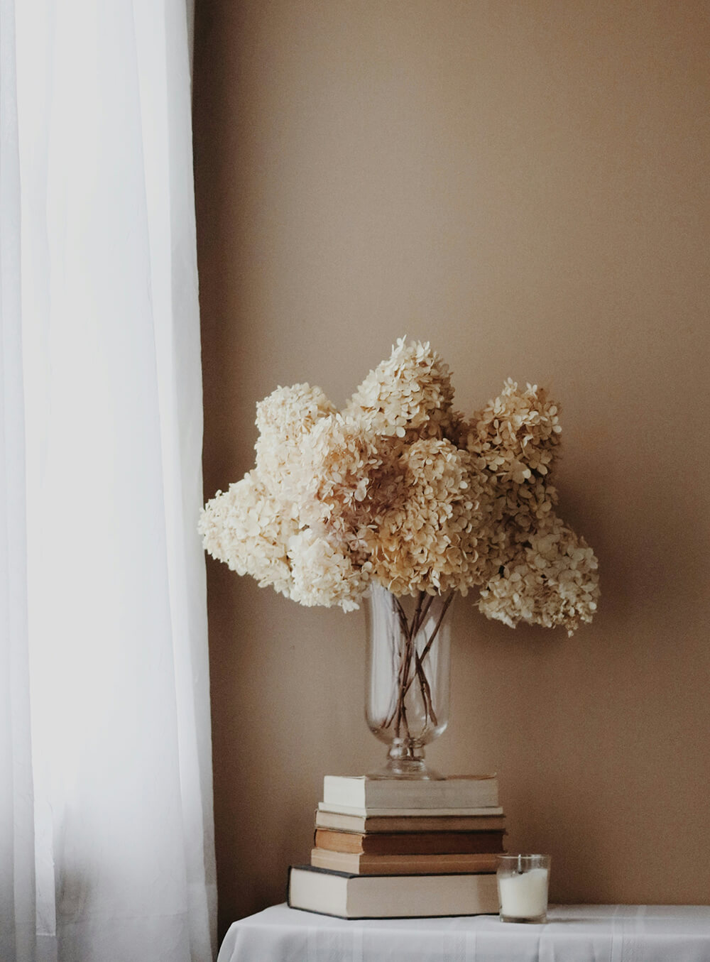 Elegant floral arrangement of dried hydrangeas on a side table with books and a candle in a minimalist home setting.