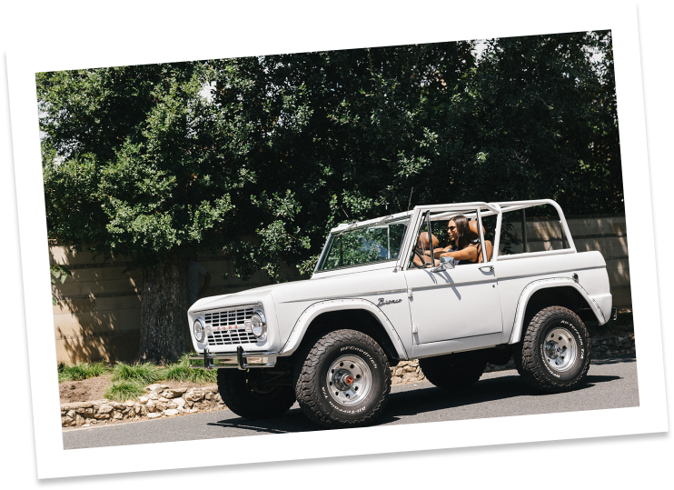 Old white For Bronco without a roof and woman driving down the street on a sunny day.